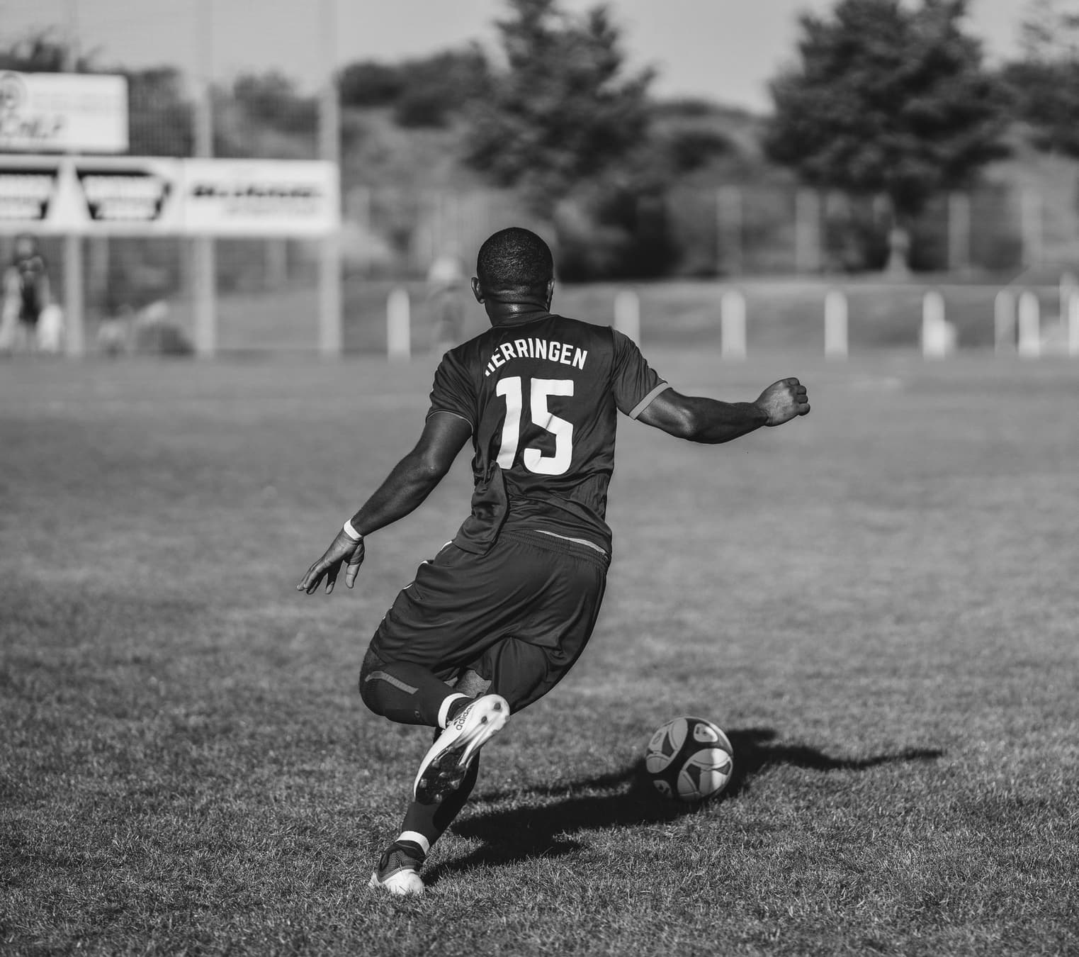 A footballer seen from behind, drawing his boot back to strike a ball on a grass pitch.